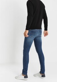 Hombre vistiendo jeans ajustados azules oscuros, camiseta de manga larga negra y zapatillas blancas, de pie contra un fondo blanco liso.