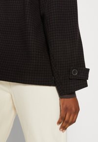 Manche de veste marron foncé à motif pied-de-poule avec poignet boutonné sur pantalon crème, montrant la main d’une personne reposant à côté de sa jambe.