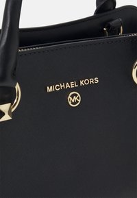 Sort læderhåndtaske med guldhardware, Michael Kors-logo og initialer nær toplynlås og håndtagsforbindelser.