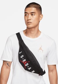 Mala cruzada preta com a palavra "JORDAN" em branco e vermelho, apresentando um logótipo. Feita de um tecido suave, usada sobre uma camisa branca.