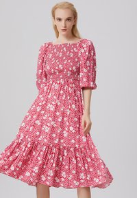 Robe rouge à fleurs avec des manches bouffantes et un corsage smocké. Elle présente une jupe à volants en étages, fabriquée à partir d'un tissu léger et fluide.