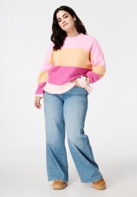 Pull en tricot coloré à rayures horizontales roses, orange et violettes. Porté avec un jean évasé bleu clair et des bottes marron clair.