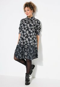Abito camicia nero con un motivo geometrico bianco, abbottonatura frontale e maniche arrotolate, abbinato a stivali neri alla caviglia e collant.