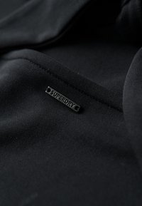 Czarna tkanina z bliska z przeszytym szwem i małą prostokątną metalową metką oznaczoną "Superdry".