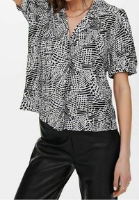 Blouse noire et blanche à manches courtes avec un motif abstrait, dotée d'un col et d'une coupe décontractée, portée avec un pantalon en cuir noir.