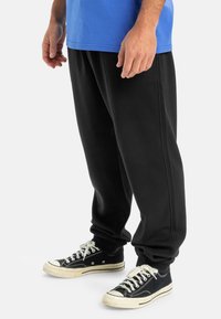 Pantalones de chándal negros con costuras laterales, puños acanalados y un ajuste relajado, combinados con zapatillas de lona en blanco y negro. Camiseta azul encima.