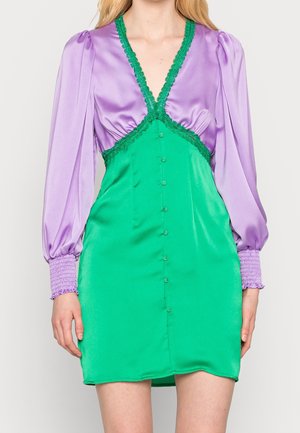 Robe avec manches longues bouffantes lavande et poignets smockés, jupe boutonnée verte, et encolure en V verte ornée de dentelle reliant le corsage et la jupe.