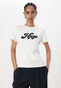 T-shirt en coton blanc avec un grand logo "Hugo" noir. Design simple avec un col rond et des manches courtes, associé à un pantalon foncé.