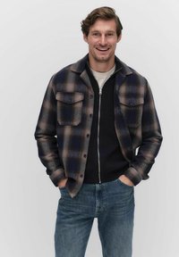 Marinblå och brun rutig flanellskjorta med två bröstfickor, bärs över en svart zip-up tröja, ihop med blåjeans.