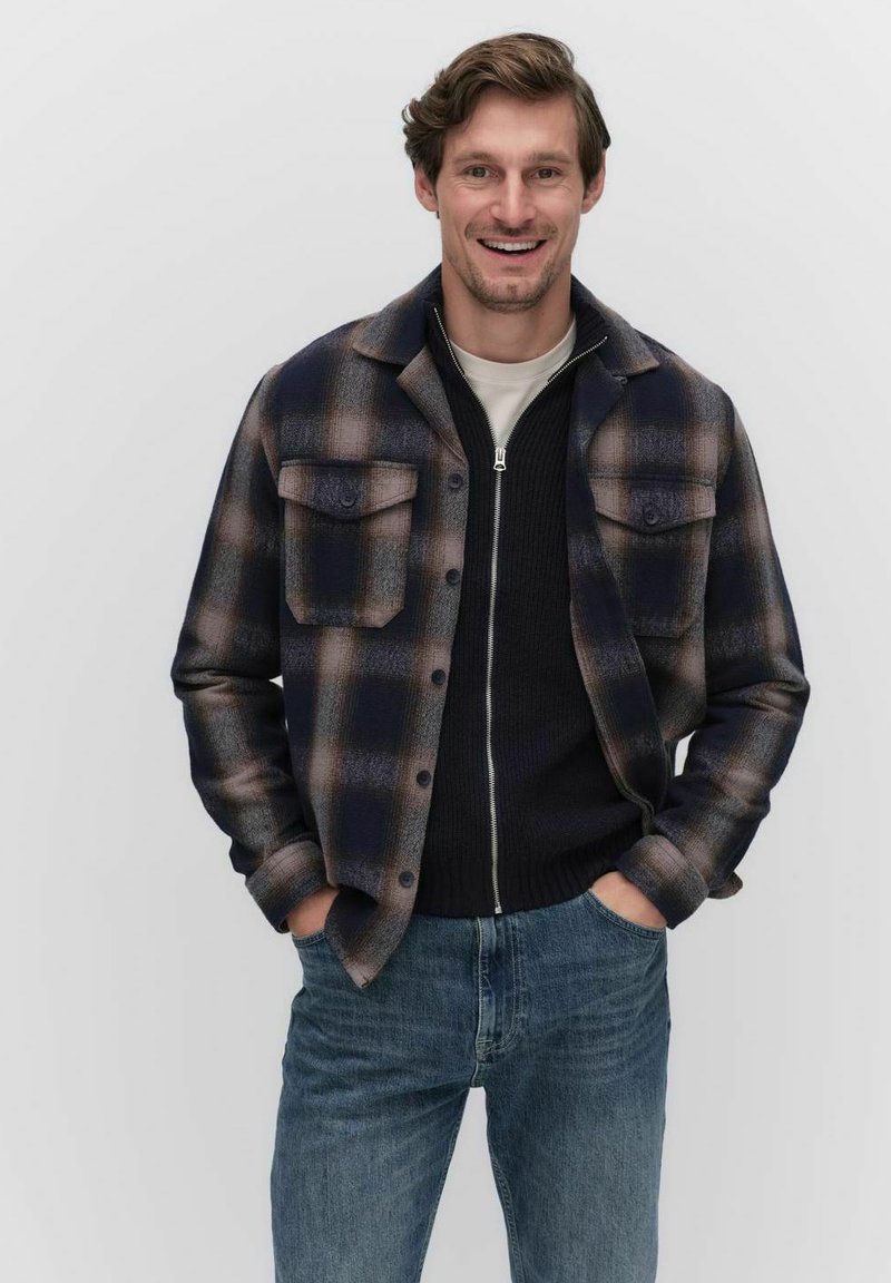 Marinblå och brun rutig flanellskjorta med två bröstfickor, bärs över en svart zip-up tröja, ihop med blåjeans.