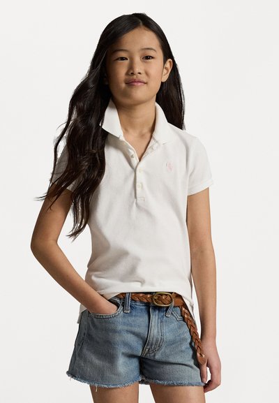 Λευκό κοντομάνικο πουκάμισο με αναδιπλωμένο γιακά, ροζ λεπτομέρεια λογότυπου στο στήθος, συνδυασμένο με μπλε τζην σορτς και καφέ πλεχτή ζώνη.