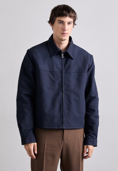 Veste bleu marine zippée à l'avant avec col et épaules structurées. Fabriquée en tissu lisse, elle présente une couture sur la poitrine et des poches latérales.