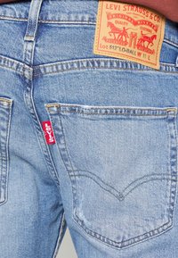 Vaqueros de mezclilla azul claro con un acabado desgastado, con una etiqueta roja de Levi's y una etiqueta marrón. Costuras visibles y diseño clásico de bolsillo trasero.