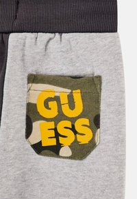 Szare i czarne dresy mają kieszeń w kamuflażu z żółtym napisem "GUESS". Materiał jest miękki, z prążkowanym pasem i teksturą po bokach.
