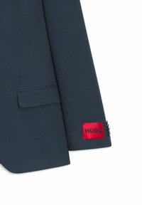 Blazer blu scuro con tessuto strutturato, tasca quadrata e etichetta rossa con la scritta "HUGO" sulla manica. Presenta dettagli con bottoni.