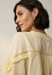 Jasnożółta bluzka z koronkowymi akcentami, falbaniaste rękawy i luźny krój. Zawiera delikatną lamówkę wokół dekoltu i ramion.