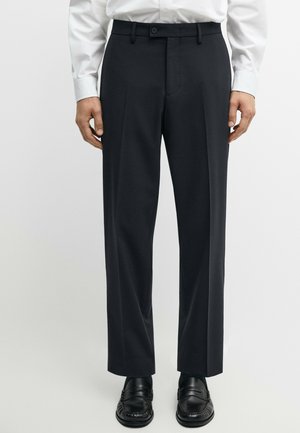 Mężczyzna ubrany w czarne, szyte na miarę spodnie, czarne eleganckie buty oraz białą koszulę z długim rękawem zapinaną na guziki, stojący na jednolitym tle.