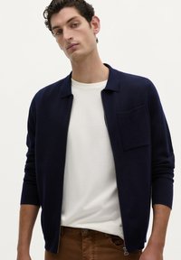 Mørkeblå zip-up cardigan lavet af tekstureret stof, med krave og en brystlomme, båret over en hvid t-shirt og brune bukser.