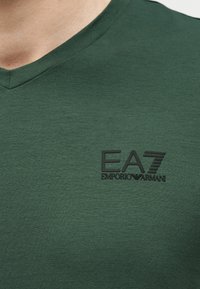 Κοντινή λήψη πράσινου μπλουζιού με V-λαιμόκοψη και μαύρο λογότυπο EA7 Emporio Armani στο αριστερό στήθος.