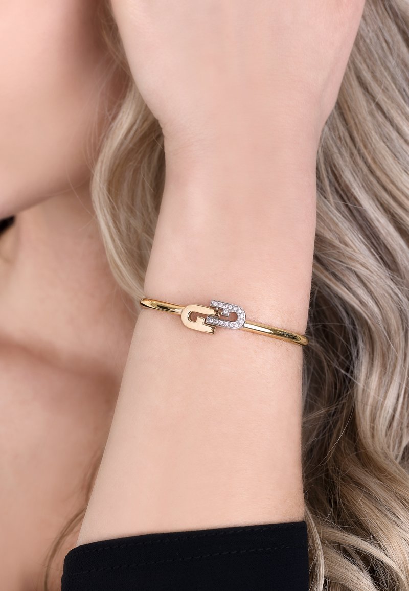 Bracciale rigido in oro con design a loop e un accento di zirconia cubica all'apertura. Finitura liscia e lucida con una forma elegante.
