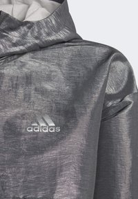 Šedá větrovka s lesklým, strukturovaným povrchem, vysokým límcem a stříbrným logem Adidas na hrudi. Lehký design.