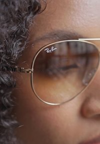 Ochelari de soare aviator cu un gradient de nuanțe maro, având un cadru subțire din aur. Prezintă logo-ul Ray-Ban pe lentilă și reflectă un fundal neclar.