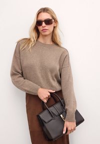 Maglione marrone a maglia con polsini a costine, abbinato a una gonna in suede marrone. Tiene una borsa a tracolla nera in pelle con dettaglio in oro sulla chiusura.