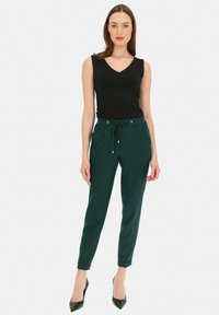 Czarny bezrękawnik z dekoltem w serek, w połączeniu z zielonymi spodniami nawiązującymi do sznurkowego zapięcia, z srebrnymi detalami, slim fit i zwężanymi kostkami. Szpilki.