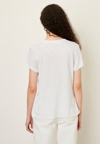 Balta trumpa rankovėmis marškinėliai iš lengvos, šiek tiek tekstūruotos medžiagos, su apvaliu kaklu ir lenktu apačiu, matomomis siūlių detalėmis.