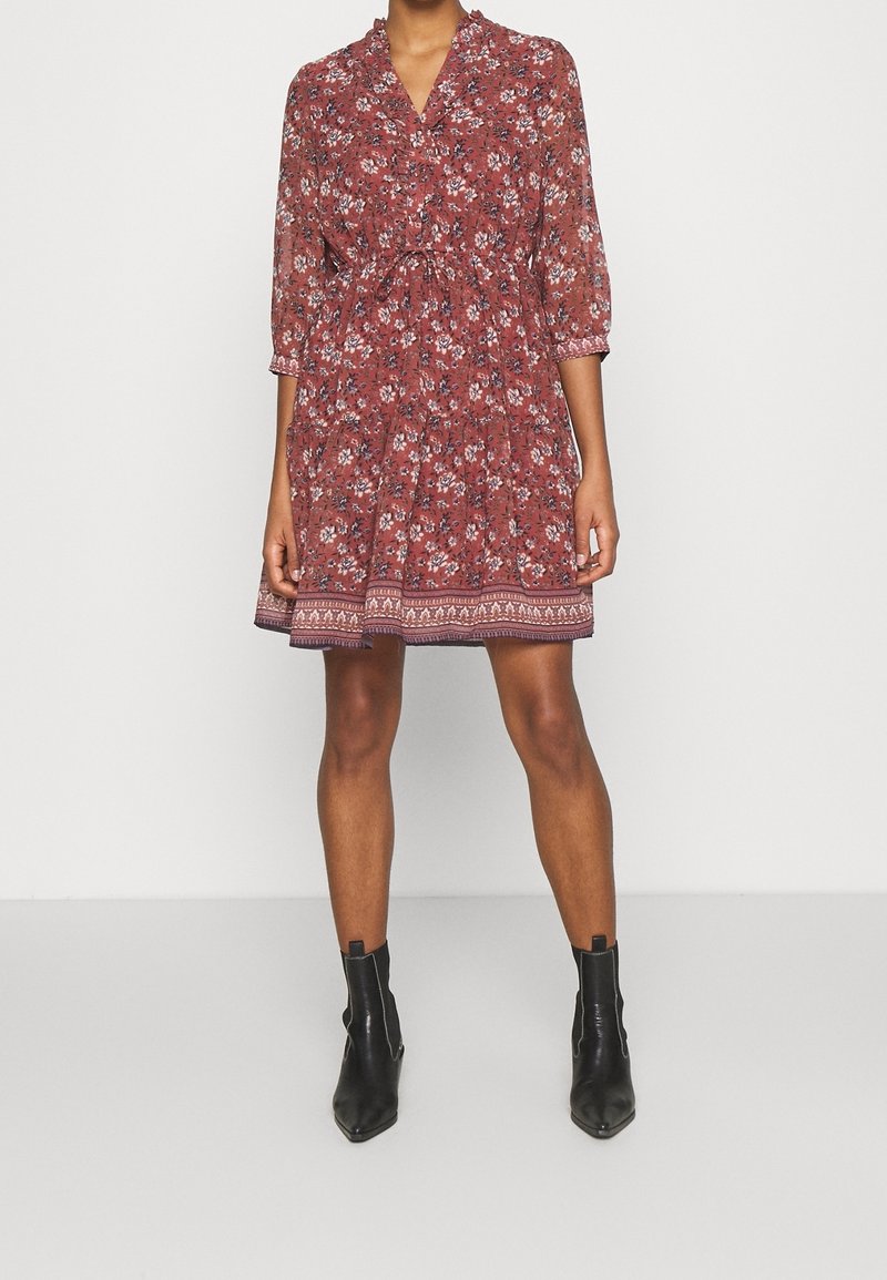 Femme portant une robe florale rouille à manches trois-quarts et des bottines noires, debout devant un fond clair uni.