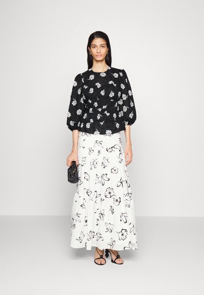 Blusa preta com padrão floral e mangas bufantes, combinada com uma saia maxi branca com padrões florais pretos. O modelo segura uma pequena bolsa preta.