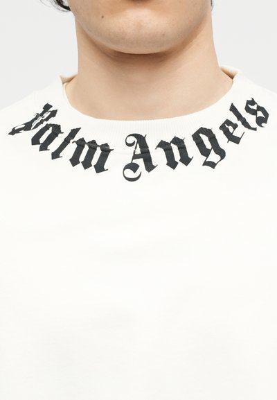 Κοντινό πλάνο ενός ατόμου που φοράει λευκό μπλουζάκι με στρογγυλή λαιμόκοψη, με τη φράση «Palm Angels» σε μαύρη γοτθική γραμματοσειρά να καμπυλώνει κατά μήκος της λαιμόκοψης.