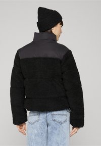 Osoba nosící černou čepici, černou fleecovou bundu s vycpanými rameny a světle modré džínové kalhoty, otočená zády k jednoduchému pozadí.