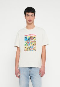 Mladić u bijeloj majici s šarenim strip panelima superheroja DC Comicsa i svijetloplavim trapericama, na bijeloj jednobojnoj pozadini.