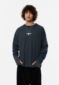 T-shirt a maniche lunghe grigio scuro con un logo bianco sul petto, caratterizzato da polsini grigi a contrasto e una vestibilità rilassata. Indossato con pantaloni in velluto a coste neri.