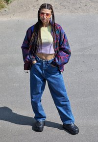 Colorful patterned jacket, yellow and purple cropped top, loose blue jeans, black clogs, braided hair, standing on a textured surface.