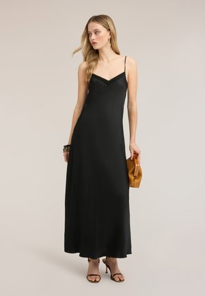Frau trägt ein langes schwarzes Slip-Kleid mit dünnen Trägern, hält eine braune Clutch, trägt Sandalen mit Absatz und goldene Ohrringe.