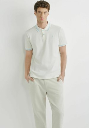 Mężczyzna ubrany w biały polo z jasnoniebieskim wykończeniem i beżowe spodnie, stojący na tle jednolitej białej ściany, z rękami w kieszeniach.