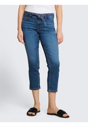 Vrouw draagt mid-rise cropped blauwe spijkerbroeken met een trekkoord in de taille en zwarte open-toe instapsandalen, staand tegen een lichte achtergrond.