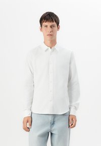 Joven de pie, vistiendo una camisa blanca de botones y jeans azul claro, contra un fondo blanco liso.