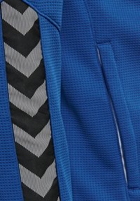 Blå texturerad jacka med panel i svart och vitt zigzag-mönster. Har sidofickor och en andningsbar design.