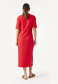 Vestido de algodón rojo con mangas cortas, aberturas laterales y un diseño recto, que llega a media pantorrilla, combinado con sandalias planas de color beige.