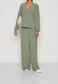 Ljust grön, strukturerad, omslagstopp med långa puffärmar, parat med matchande vida byxor. Accessoarer i form av en silverfärgad franshandväska.