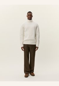Ribbed turtleneck sweater in off-white, paired with brown checkered pants and brown shoes. Model stands against a neutral background.