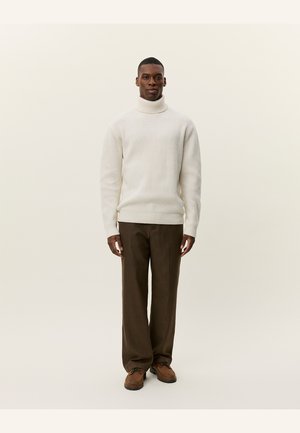 Ribbed turtleneck sweater in off-white, paired with brown checkered pants and brown shoes. Model stands against a neutral background.