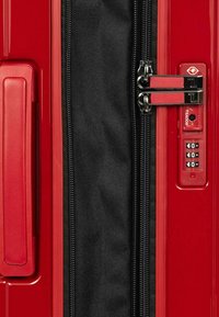 Valigia rigida rossa con superficie texturizzata, dotata di doppie zip nere, lucchetto TSA integrato e maniglia laterale.