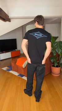 Άνδρας με καστανά μαλλιά που φοράει μαύρο μπλουζάκι Rip Curl και σκούρο τζιν, στέκεται σε ένα σαλόνι με ξύλινο πάτωμα και πορτοκαλί καναπέ.