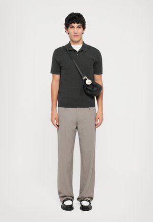 Polo shirt nero a maniche corte, pantaloni beige, borsa nera con dettagli dorati e scarpe nere con accenti bianchi. Sfondo semplice.