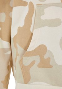 Beige kamuflasjehette med lomme, laget av mykt stoff. Har bølgende mønstre i lysebeige og kremfarger, med synlig søm.