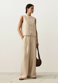 Femme debout portant un haut sans manches beige texturé avec des boutons sur le devant et un pantalon large assorti, tenant un sac marron à ses côtés.